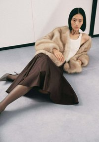 Abrigo de piel beige, camiseta sin mangas blanca, falda marrón fluida, medias transparentes y tacones grises. La modelo posó sobre un suelo gris claro con fondo minimalista.