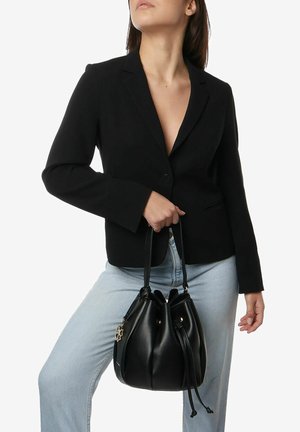 Borsa a secchiello in pelle nera con chiusura a coulisse, texture liscia e hardware decorativo. Indossata con un blazer nero e jeans azzurri.