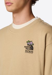 Beige sweatshirt med rund halsringning, ribbad halslinning och flerfärgad broderad logotyp med blomsterdetaljer på vänster bröst.