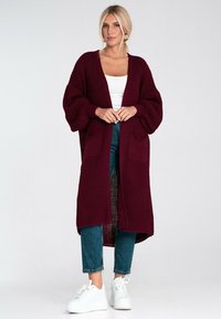 Burgundowy sweter z dzianiny o prążkowanej teksturze, z otwartym przodem, oversize'owymi rękawami i dwiema kieszeniami z przodu, zestawiony z dżinsami i białymi trampkami.