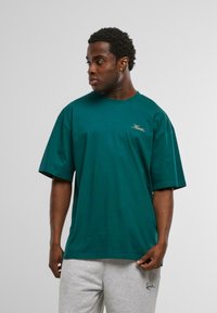 Tricou din bumbac de culoare verde închis, cu croială lejeră, mâneci scurte și un mic logo brodat cu fir auriu pe piept. Este purtat împreună cu pantaloni de trening gri.