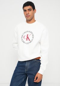 Λευκό φούτερ με στρογγυλή λαιμόκοψη, διακοσμημένο με κυκλικό κόκκινο και γκρι γραφικό λογότυπο. Φορεμένο με μπλε τζιν. Απαλή υφή και άνετη εφαρμογή.