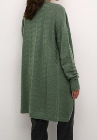 Sweter kardigan w jasnozielonym kolorze, z teksturowanym, zygzakowatym wzorem, długimi rękawami i luźnym krojem, który opada poniżej bioder.