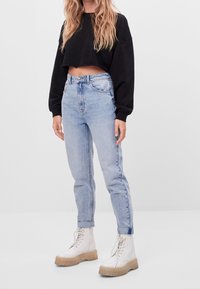 Svart croppad sweatshirt, ljusblå hög midja jeans och vita snörstövlar med en tjock tan-sula. Enfärgad bakgrund.