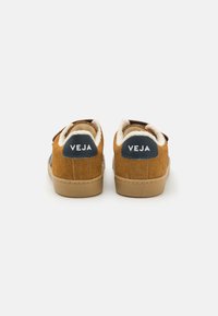 Zapatos de ante marrón con un interior acolchado, que presentan una etiqueta con el logo en azul marino en el talón y una suela de goma beige. Diseño simple, perfil bajo.