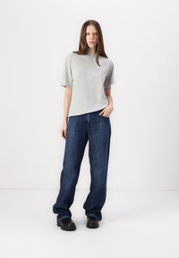Grå t-skjorte med korte ermer sammen med løse, mørkeblå denimjeans og svarte runde sko på en ensfarget bakgrunn.