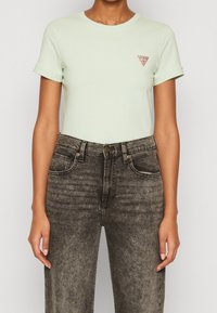 Mátová krátká trička s krátkým rukávem a malým červeným trojúhelníkovým logem na hrudi, v kombinaci s černými džínami se zvýšeným pasem a vybledlým vzhledem.