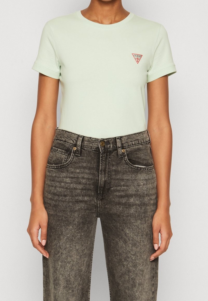 Mátová krátká trička s krátkým rukávem a malým červeným trojúhelníkovým logem na hrudi, v kombinaci s černými džínami se zvýšeným pasem a vybledlým vzhledem.