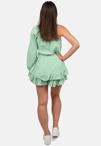 Lichtgroene jurk met één schouder, met volledig gerimpelde zoom en textuur. Model draagt witte sneakers, met los haar, vanuit de achterkant bekeken.