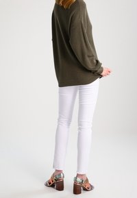 Maglione oversize verde oliva, jeans slim-fit bianchi e tacchi a blocco multicolori con cinturino trasparente e tacco in legno.