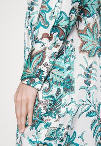 Bluzka z długim rękawem z jedwabistego materiału, z turkusowymi, brązowymi i białymi wzorami kwiatowymi. Zapięcie przy mankietach z ozdobnym ściegiem.