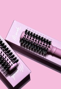 Brosse à cheveux ronde avec des poils noirs, un manche rose clair et un emballage rose comportant du texte blanc. Les poils ont des pointes métalliques et des longueurs variées.