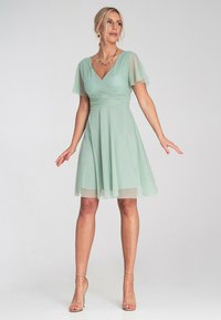 Hellgrünes Chiffonkleid mit kurzen, flatternden Ärmeln, V-Ausschnitt, geraffter Taille und einem fließenden Rock. Getragen mit nude-farbenen Riemchen-Heels.