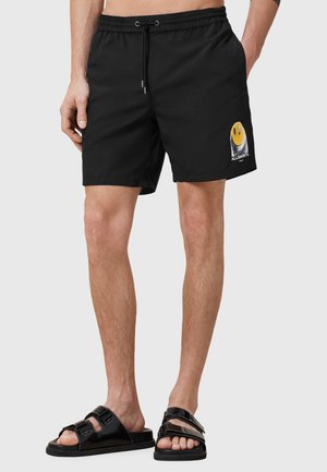 Pantaloni da bagno neri con cintura elastica e cordino, caratterizzati da una grafica di un sorriso giallo sulla gamba sinistra e una texture liscia.