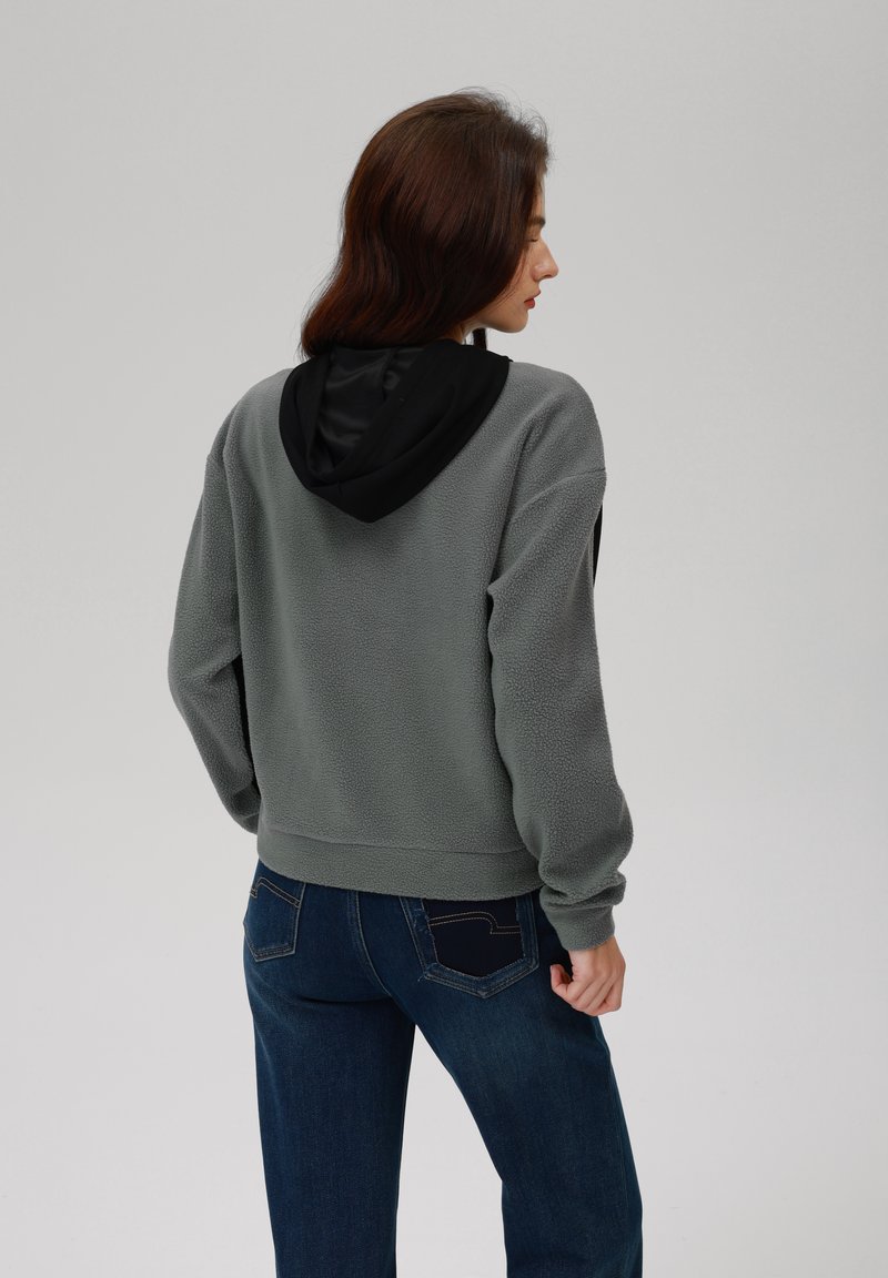 Szara polarowa bluza z czarnym kapturem, o kroju cropped i elastycznych mankietach. Zestawiona z ciemnoniebieskimi dżinsami z detalami kieszeni.