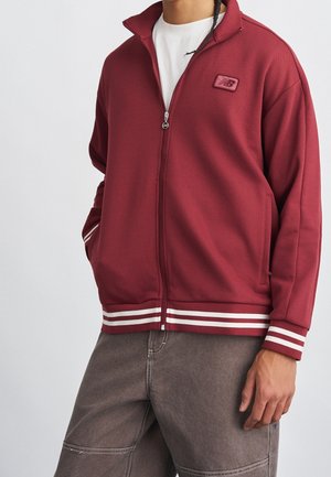 Personne portant une veste zippée bordeaux avec des poignets et un ourlet rayés blancs, associée à un pantalon gris, debout devant un fond uni.