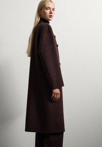 Blonde woman wearing a long dark brown coat and black turtleneck, standing sideways against a plain light gray wall.