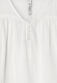 Blusa bianca, ampia, con scollo a V, chiusura con sei piccoli bottoni rotondi e tessuto morbido e strutturato.