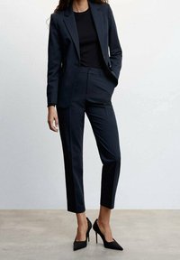 Blazer noir sur mesure et pantalon court assorti, tous deux en tissu lisse. Associés à des escarpins noirs à talons aiguilles, créant un look costume formel.