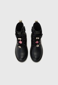 Bottines en cuir noir avec des accessoires en or, ornées de charms décoratifs incluant un cœur rose et une étiquette 9¾, doublure intérieure texturée.