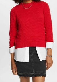Pull rouge en tricot avec un motif texturé, col rond, et manches trois-quarts, porté sur une chemise blanche à col et une jupe en jean noire.