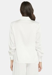 Blusa bianca a maniche lunghe con una texture liscia, caratterizzata da un sottile motivo a pois e colletto classico, con polsini visibili e bottone.