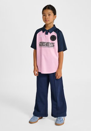 Ung pige stående fremadrettet iført en pink og navy sportstrøje, navy vidde bukser og lyseblå sneakers.