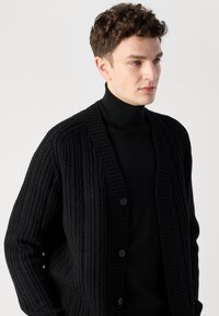 Μαύρο κασκόλ με ριγέ υφή, με κουμπιά μπροστά και μακριά μανίκια, σε στρώσεις πάνω από ένα μαύρο ζιβάγκο. Μινιμαλιστικός σχεδιασμός.