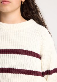 Nainen, jolla on päällään kerma- värinen neulepusero, jossa on kaksi vaakasuoraa tummanpunaiseta raitaa, ja kullasta valmistettu renkaankorvake, jossa on pieni riipus. Hänen pitkät tummat kiharat hiuksensa ovat näkyvissä.