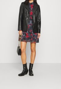 Blazer de cuero negro sobre un vestido floral en tonos azul oscuro y rojo. La modelo lleva botas negras hasta el tobillo y sostiene un bolso negro con una correa de cadena.