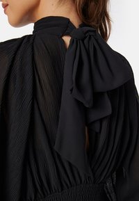 Blouse noire avec une texture plissée, un col haut et un détail de nœud noué dans le dos. Le tissu semble léger et fluide.