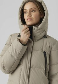Chaqueta acolchada beige con capucha, cuenta con un bolsillo en el pecho con cremallera, correas ajustables y una textura suave y mate.