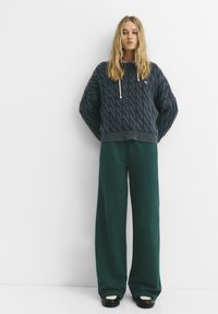 Dundi szürke kötött pulóver fehér húzózsinórral, laza sötétzöld nadrággal és fekete sneakerekkel párosítva. Egyszerű és texturált outfit.