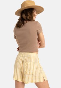 Top scurt, ajustat, de culoare maro, asortat cu pantaloni scurți cu dungi galbene, având o talie elastică și buzunare laterale. Purtați cu o pălărie de paie cu boruri late.