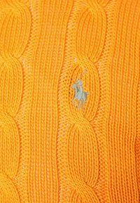 Gros plan sur un tissu en maille torsadée orange vif avec une petite figure brodée représentant une personne à cheval tenant un maillet.
