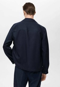 Marineblaue Jacke mit geradem Schnitt, langen Ärmeln und spitzem Kragen. Aus einem glatten, robusten Stoff gefertigt, mit minimalen Details.