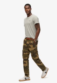 Man walking, wearing a light gray short-sleeve t-shirt, brown camouflage cargo pants, white sneakers, and a brown belt with a watch.