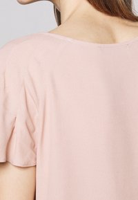 Jasnoróżowa bluzka z miękkiego, teksturowanego materiału, z szerokim dekoltem i krótkimi rękawami. Krój jest luźny w okolicy ramion.
