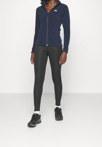 Chaqueta de forro polar de color navy con cierre de cremallera y cuello alto, combinada con leggings negros y zapatillas deportivas negras. Logotipo blanco en la chaqueta.