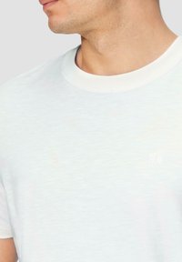 Weißes Kurzarm-T-Shirt mit rundem Ausschnitt, das ein dezentes Logodetail und einen weichen, strukturierten Stoff aufweist.
