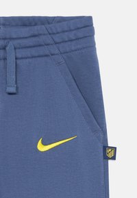 Blaue Sweatpants aus Baumwollmischung, mit einem gelben Nike-Logo, seitlichen Taschen und einem Atlético Madrid-Label an der Seite. Glatte Textur.