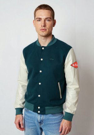 Junger Mann steht, trägt eine türkis-weiße Collegejacke mit Knöpfen und hellblaue Jeans, blickt nach vorne vor einem einfarbigen Hintergrund.