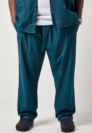 Ein Mann steht mit den Händen in den Taschen und trägt eine türkisfarbene Leinenhose, ein passendes kurzärmeliges Hemd, ein weißes Unterhemd, schwarze Schuhe und eine silberne Armbanduhr.