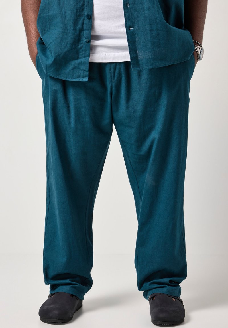 Un uomo in piedi con le mani in tasca indossa pantaloni di lino turchese, camicia a maniche corte coordinata, maglietta bianca sotto, scarpe nere e orologio da polso argentato.