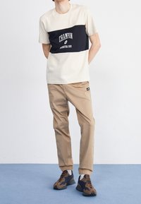 Krämfärgad och svart t-shirt med kort ärm och "Champion"-logga, beige joggerbyxor och bruna sneakers med marinblå detaljer.