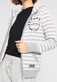 Donna con una felpa grigia e bianca a strisce con zip sopra una maglietta nera e pantaloni della tuta grigi, che tiene la zip della felpa con una mano.