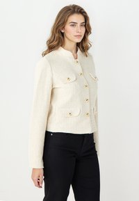 Cremefarbene Bouclé-Jacke mit rundem Ausschnitt, vier aufgesetzten Taschen vorne und goldfarbenen Knopfakzenten. Kurz geschnittenes Design, strukturierter Stoff. Kombiniert mit schwarzen Hosen.