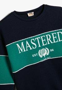 Sudadera azul marino con una franja horizontal verde que lleva la palabra "MASTERED" en texto blanco, adornada con un motivo de golf y costuras en contraste.