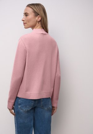 Cardigan rosa lavorato a maglia con collo alto, polsini e orlo a coste. Abbinato a jeans blu dal taglio morbido. Sottile trama sul tessuto.