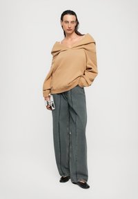 Femme portant un sweat-shirt beige oversize à épaules nues, un jean gris à jambes larges, des chaussures noires, tenant un petit sac à main blanc, se tenant sur fond blanc.
